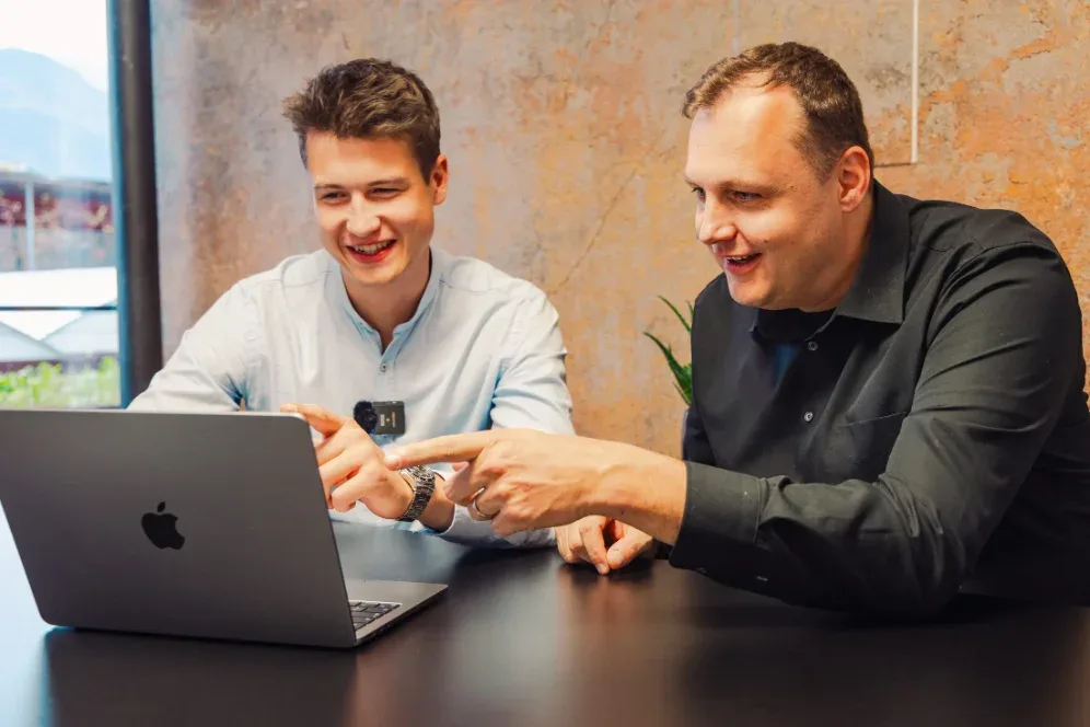 Zwei Männer sehen sich lachend eine Website am Laptop an, einer zeigt auf den Bildschirm.